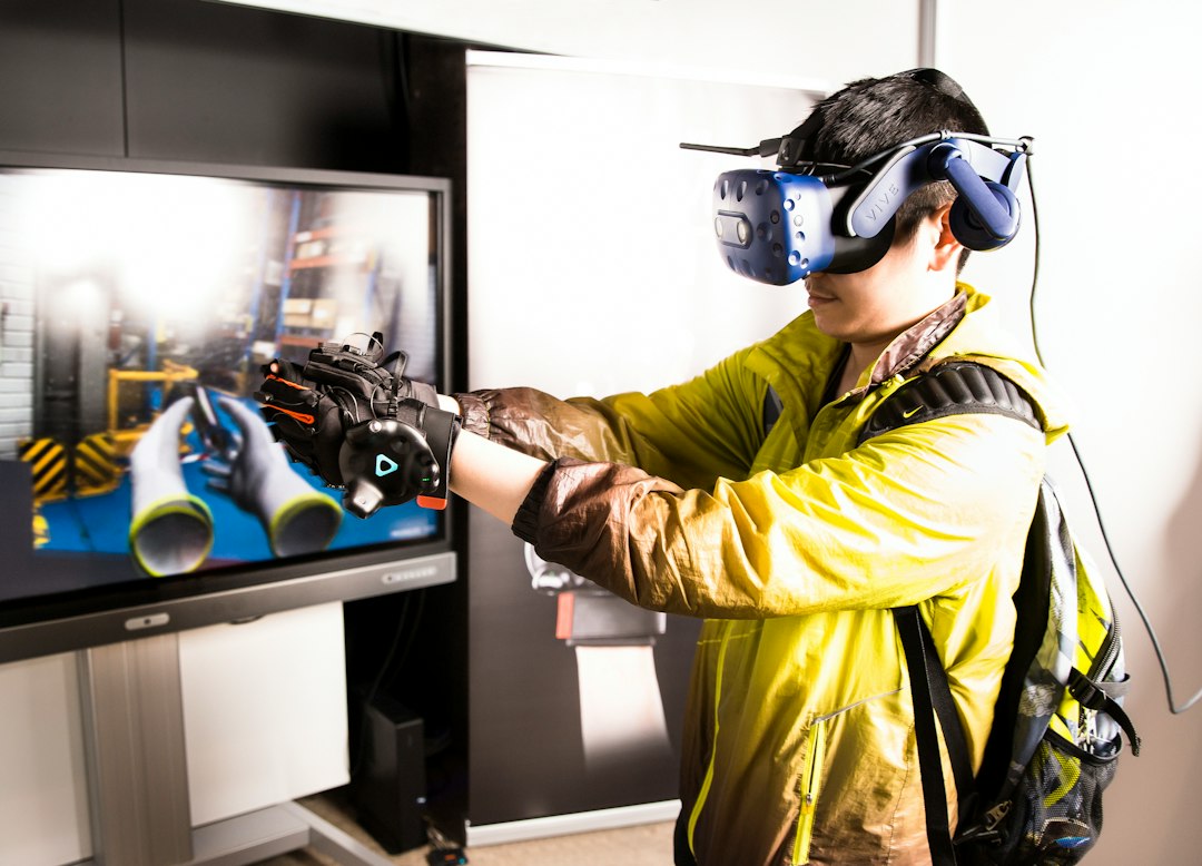Trainee in a yellow jacket wearing a VR headset and hand controllers in front of an industrial scene on screen